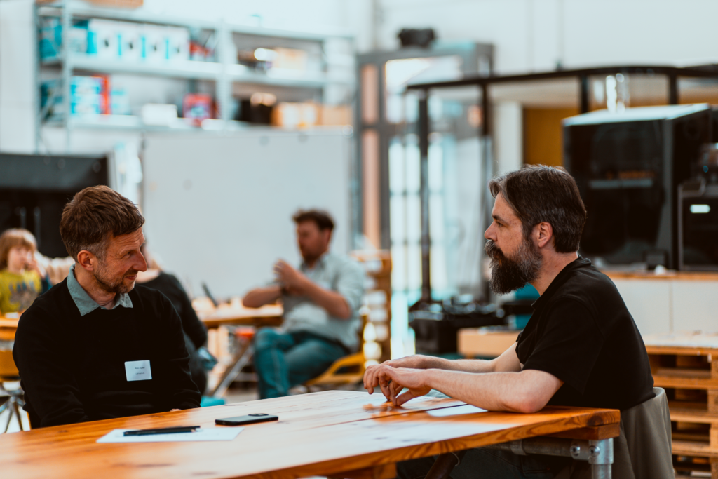 Zwei Männer sitzen sich an einem Holztisch in einem hellen, offenen Raum gegenüber und führen ein konzentriertes Gespräch; im Hintergrund arbeiten weitere Personen in einer workshopartigen Atmosphäre.