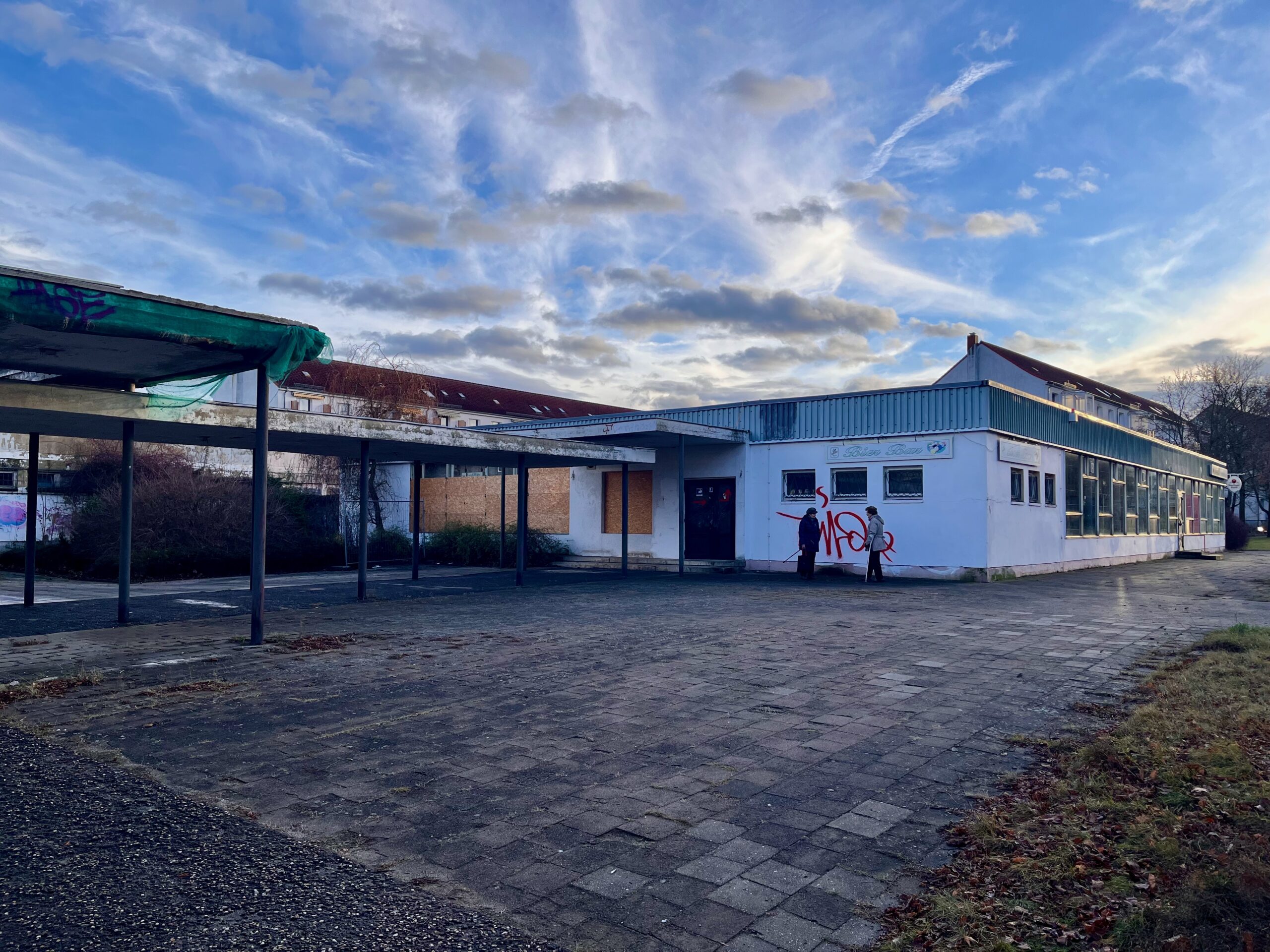 Außenaufnahme des „Club am Anger“ in Eisenhüttenstadt. Zu sehen ist ein flaches, weißes Bestandsgebäude mit blauer Fassadenverkleidung, großen Fensterfronten, teilweise verbretterten Fenstern und Graffiti an der Wand. Vor dem Gebäude liegt ein großer gepflasterter Platz mit überdachten Bereichen; zwei Personen stehen nahe dem Eingang. Darüber spannt sich ein weiter blauer Himmel mit auffälligen Wolkenformationen.