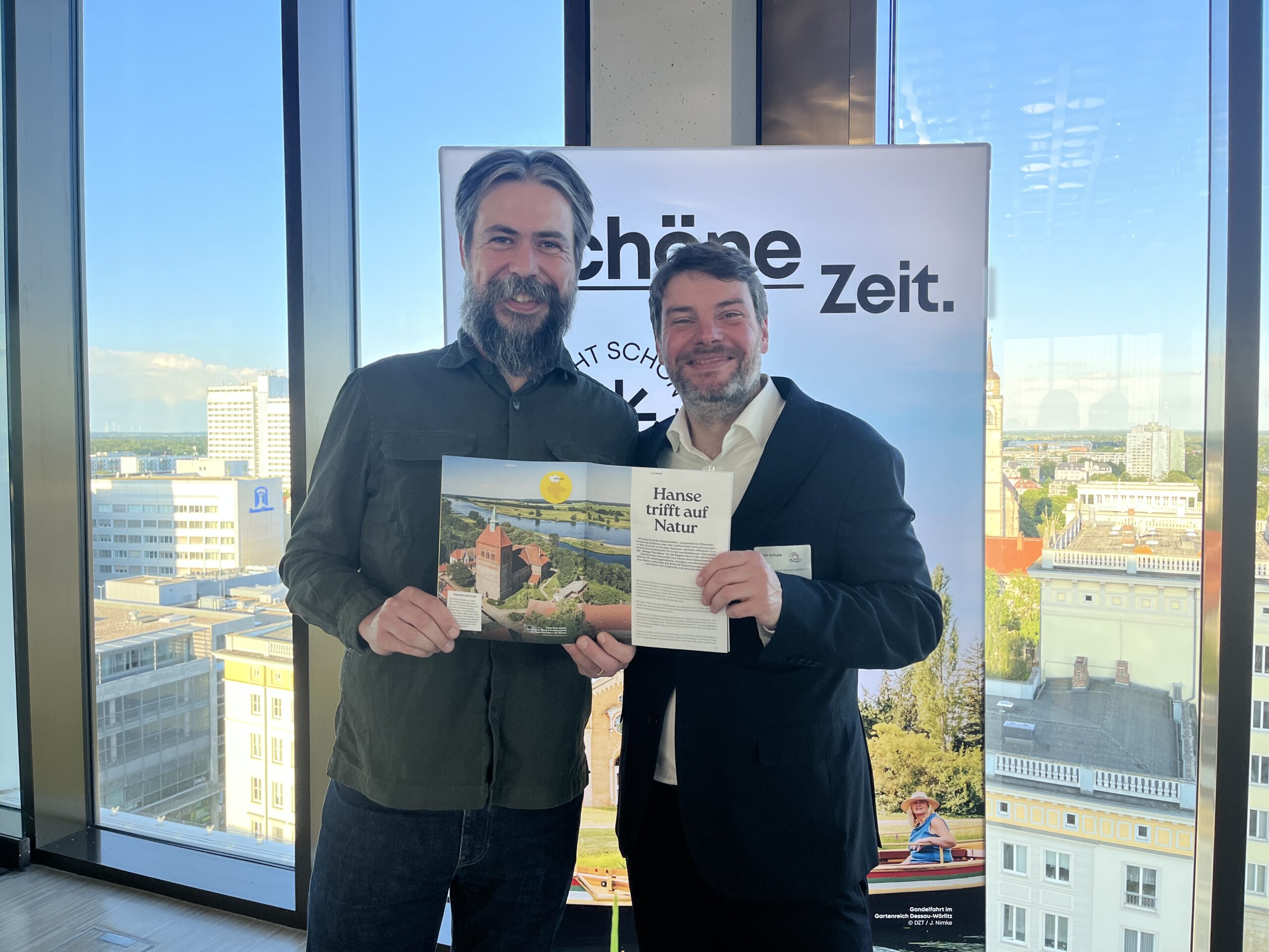 Zwei Männer stehen bei einer Veranstaltung vor großen Fenstern mit Blick über Magdeburg. Links steht Tobias Kremkau, rechts Martin Schulze, Geschäftsführer des Landestourismusverbands Sachsen-Anhalt. Beide lächeln in die Kamera und halten gemeinsam ein aufgeschlagenes Magazin mit touristischen Motiven aus Sachsen-Anhalt. Im Hintergrund ist ein Roll-up mit der Aufschrift „Schöne Zeit.“ zu sehen.