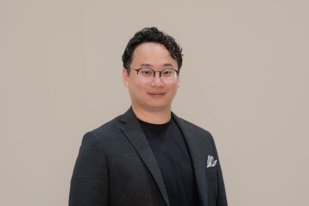 Das Porträtfoto zeigt Yuta Aoki vor schlichtem beigem Hintergrund. Er trägt eine dunkle Jacke über einem schwarzen Oberteil, eine Brille und blickt mit leichtem Lächeln direkt in die Kamera.